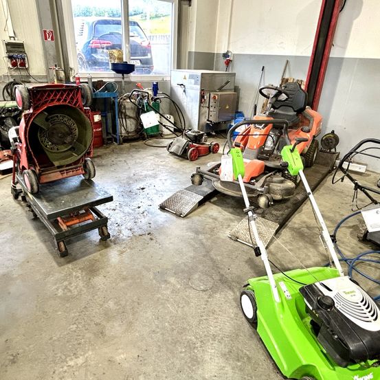 Eine Garage mit verschiedenen Rasenmähern und Ausrüstung, einschließlich eines roten Gebläses und eines orangefarbenen Rasenmähers auf einer Hebebühne.
