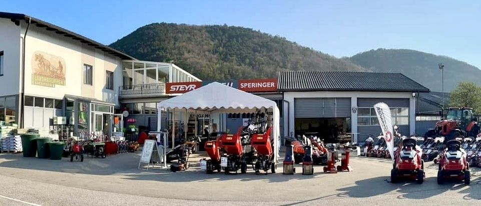 Ein Händler mit einer großen Auswahl an roter Maschinen, einschließlich Rasenmäher, geparkt vor einem Gebäude mit einem weißen Baldachin und einem Schild, das STEYR SPERINGER liest.