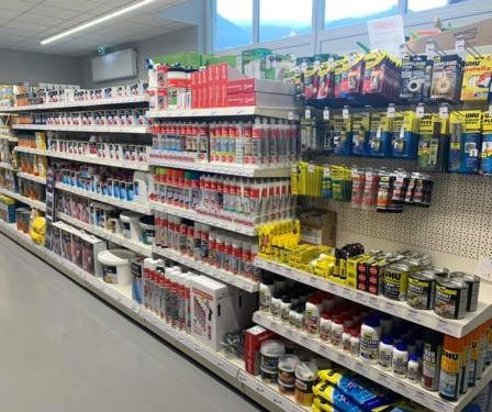 Ein Gang in einem Geschäft gefüllt mit verschiedenen Produkten. Die Regale sind mit Flaschen, Dosen und Kartons gefüllt, alle beschriftet. Der Boden ist grau, und die Decke hat weiße Lampen.