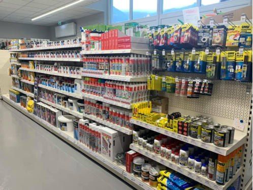 Ein Gang in einem Geschäft gefüllt mit verschiedenen Produkten. Die Regale sind mit Flaschen, Dosen und Kartons gefüllt, alle beschriftet. Der Boden ist grau, und die Decke hat weiße Lampen.