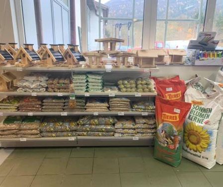 Ein Ladeninneres mit einem großen Regal, gefüllt mit Tüten Vogelfutter, Vogelhäusern und anderen Haustierbedarfsartikeln. Die Regale sind ordentlich gestapelt, und die Tüten haben farbenfrohe Etiketten.