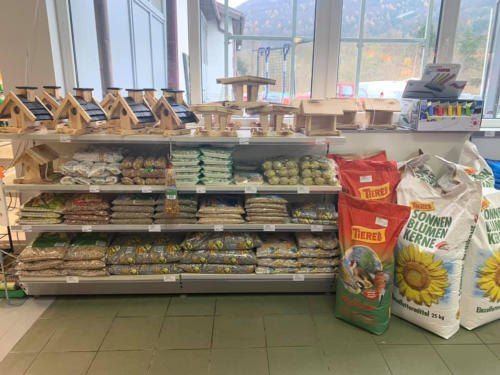 Ein Ladeninneres mit einem großen Regal, gefüllt mit Tüten Vogelfutter, Vogelhäusern und anderen Haustierbedarfsartikeln. Die Regale sind ordentlich gestapelt, und die Tüten haben farbenfrohe Etiketten.