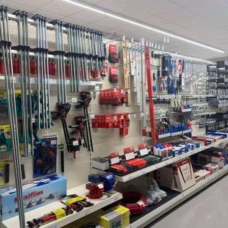 Ein Baumarkt mit verschiedenen Werkzeugen und Geräten, die ordentlich auf Regalen angeordnet sind. Jedes Regal enthält verschiedene Werkzeuge, einige hängen an Hakenbrettern. Es gibt Kisten, Behälter und Container, die Werkzeuge halten. Ein langes Regal beherbergt mehrere Werkzeuge. Eine weiße Deckenleuchte beleuchtet den Bereich.