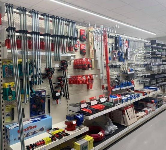 Ein Baumarkt mit verschiedenen Werkzeugen und Geräten, die ordentlich auf Regalen angeordnet sind. Jedes Regal enthält verschiedene Werkzeuge, einige hängen an Hakenbrettern. Es gibt Kisten, Behälter und Container, die Werkzeuge halten. Ein langes Regal beherbergt mehrere Werkzeuge. Eine weiße Deckenleuchte beleuchtet den Bereich.