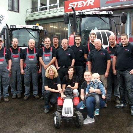 Eine Gruppe von Menschen in Arbeitskleidung steht vor roter und weißer Maschinerie. Frauen und Kinder sind ebenfalls anwesend, lächeln und posieren für ein Foto.