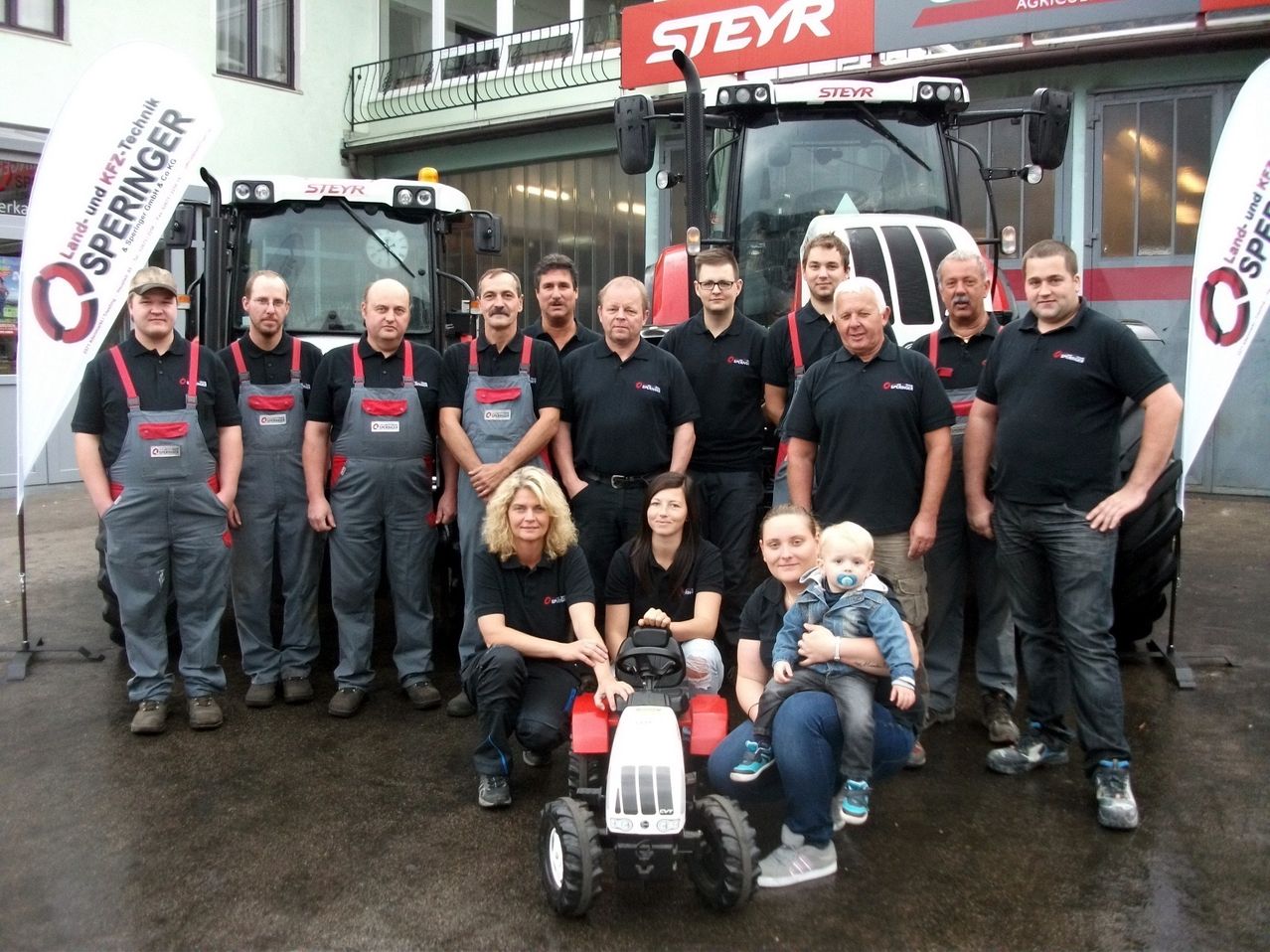 Eine Gruppe von Menschen in Arbeitskleidung steht vor roter und weißer Maschinerie. Frauen und Kinder sind ebenfalls anwesend, lächeln und posieren für ein Foto.