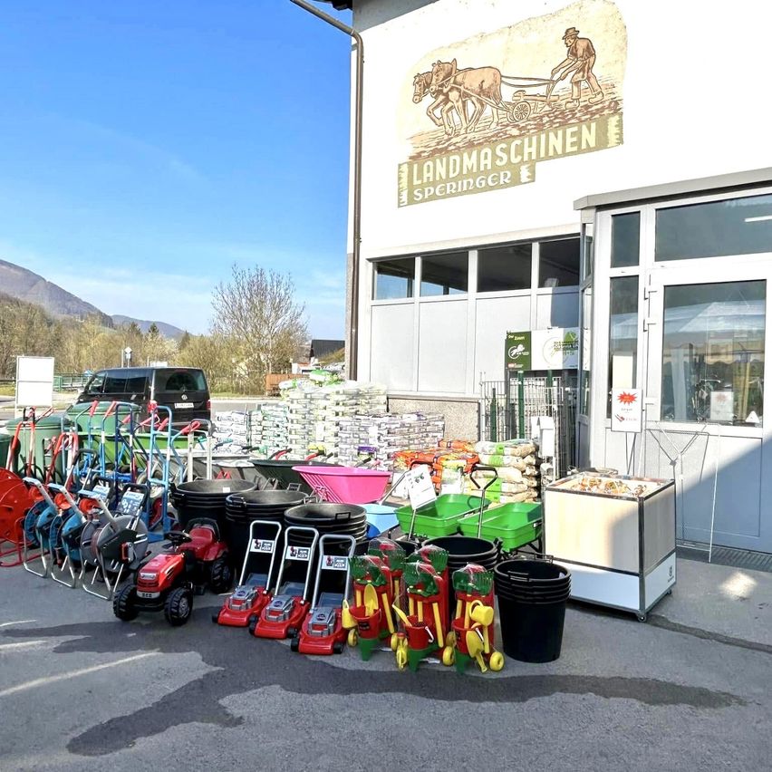 Ein Geschäft mit dem Schild 'Landmaschinen Springer' zeigt verschiedene Gartengeräte und Ausrüstung im Freien, darunter Schubkarren, Eimer und Spielzeugtraktoren.