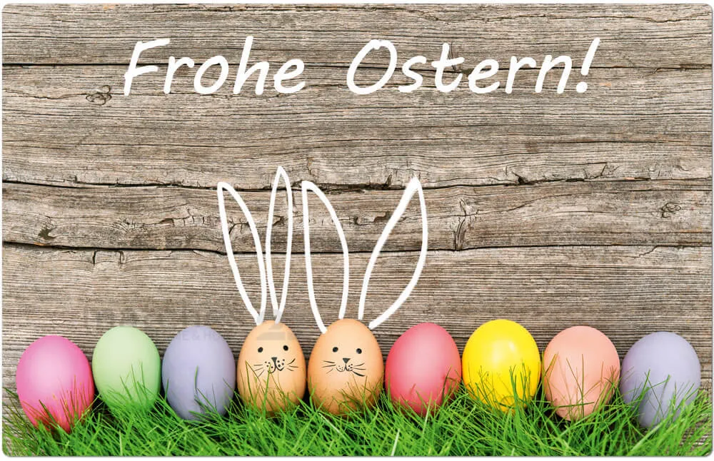 Ein hölzerner Hintergrund mit 'Frohe Ostern!' darauf geschrieben. Darunter befinden sich sieben Ostereier mit Hasenohren, die in einer Reihe auf einer grasbewachsenen Oberfläche angeordnet sind.