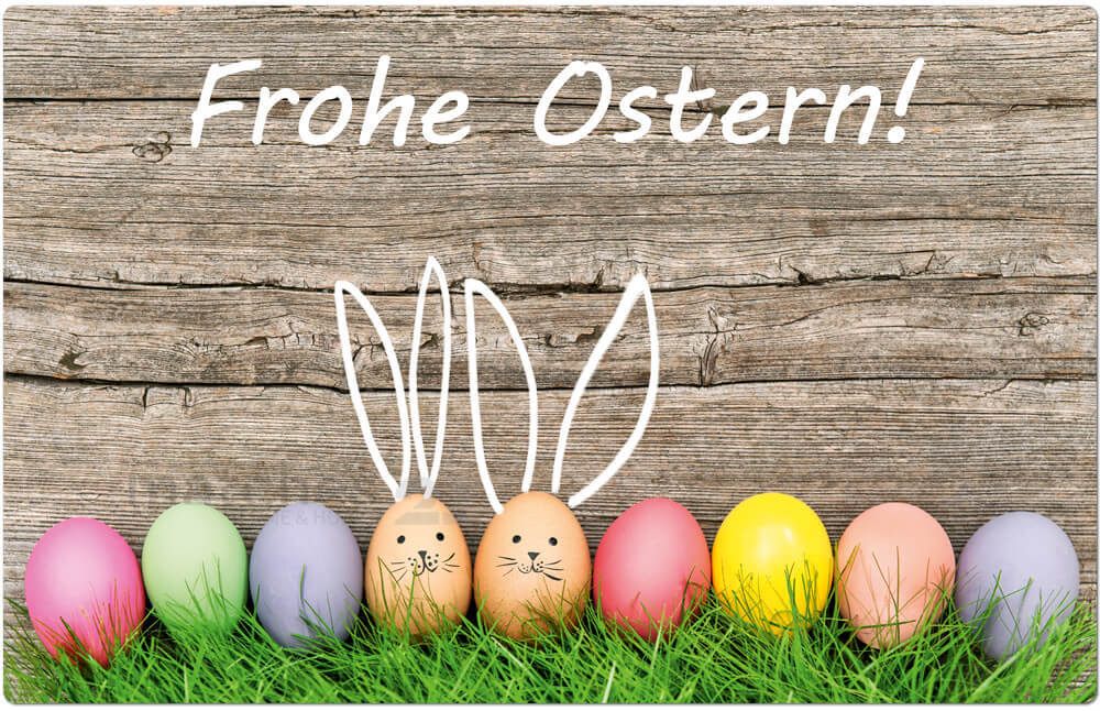 Ein hölzerner Hintergrund mit 'Frohe Ostern!' darauf geschrieben. Darunter befinden sich sieben Ostereier mit Hasenohren, die in einer Reihe auf einer grasbewachsenen Oberfläche angeordnet sind.