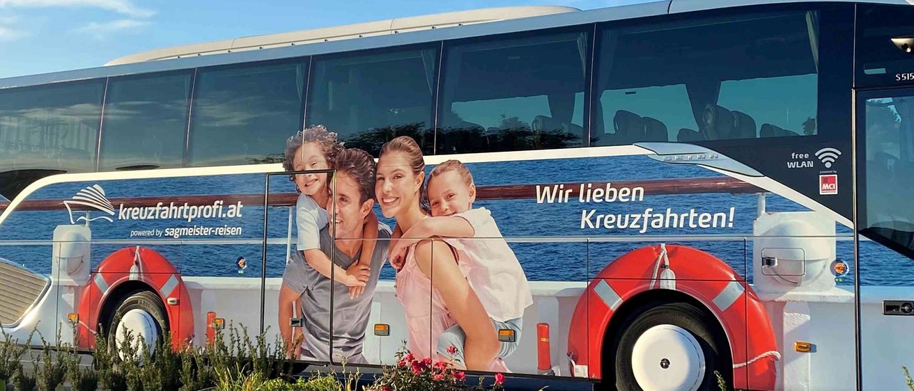 Ein großer Bus ist in einer malerischen Gegend mit Pflanzen und Blumen geparkt. Er zeigt ein Familienfoto an der Seite und wirbt für eine Kreuzfahrtfirma. Der Bus ist rot und weiß, mit Rettungsringen vorne und hinten.