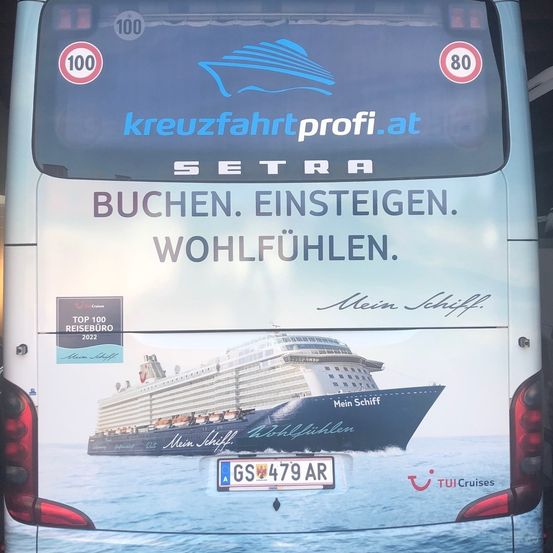 Die Rückseite eines Busses zeigt eine große Werbung für SETRA, die zum Buchen, Einsteigen und Genießen animiert. Ein Kreuzfahrtschiff ist abgebildet mit dem Text 'Mein Schiff Wohlfühlen'. Die Werbung stammt von kreuzfahrtprofi.at.