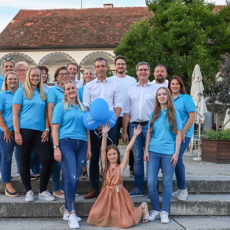 Eine Gruppe von Menschen in übereinstimmenden blauen Hemden posiert für ein Foto auf einer Treppe, mit einem Gebäude und Grünpflanzen im Hintergrund. Einige Personen halten blaue Luftballons.