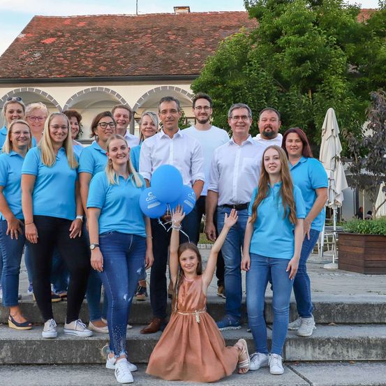Eine Gruppe von Menschen in übereinstimmenden blauen Hemden posiert für ein Foto auf einer Treppe, mit einem Gebäude und Grünpflanzen im Hintergrund. Einige Personen halten blaue Luftballons.