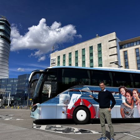 Ein Mann steht vor einem Bus am Flughafen. Der Bus hat Bilder einer Familie und eines Rettungsrings. Hinter ihm steht ein Kontrollturm und ein Hotel.