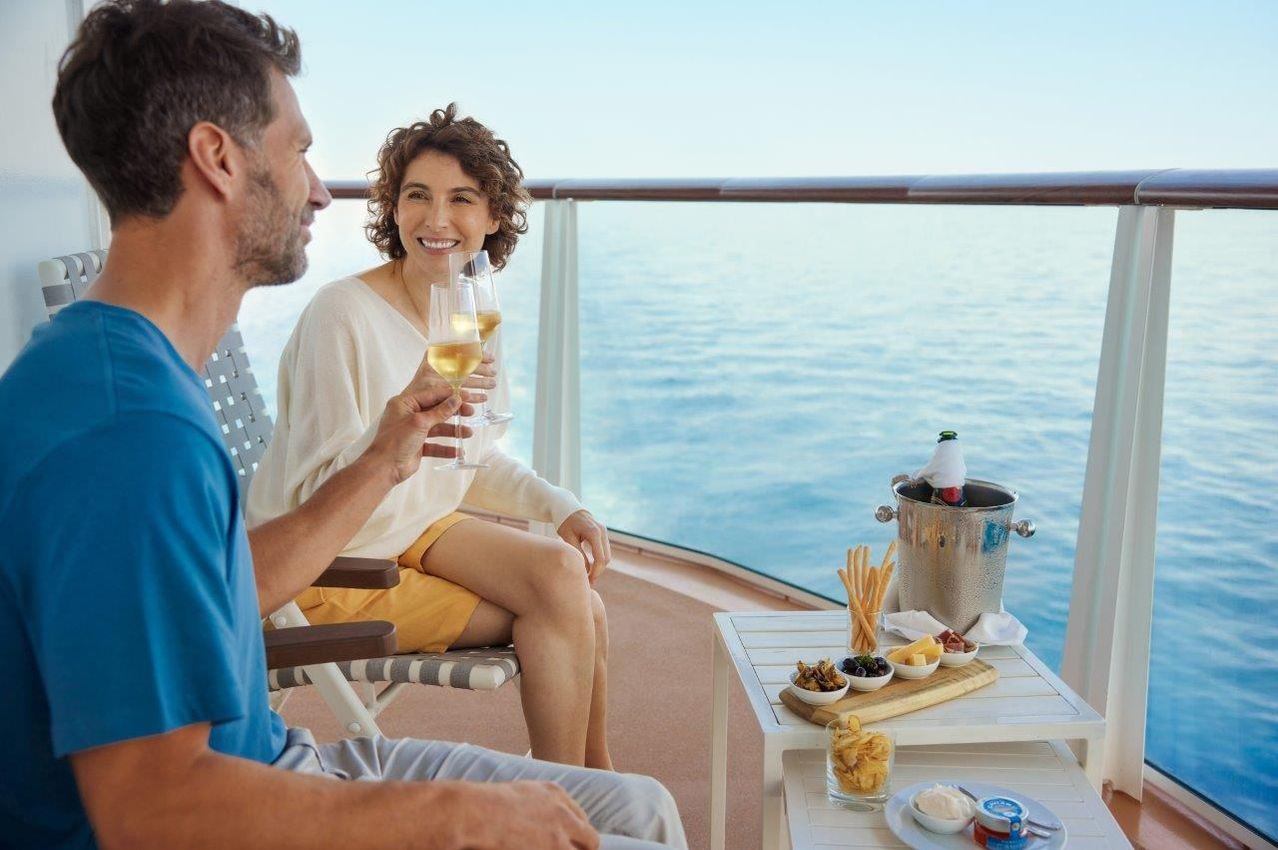 Ein Paar genießt ein Getränk auf einem Balkon mit Blick auf den Ozean. Sie sitzen auf einem Stuhl mit einem Tisch davor, der mit Snacks gefüllt ist und einer Flasche Wein in einem Eimer.