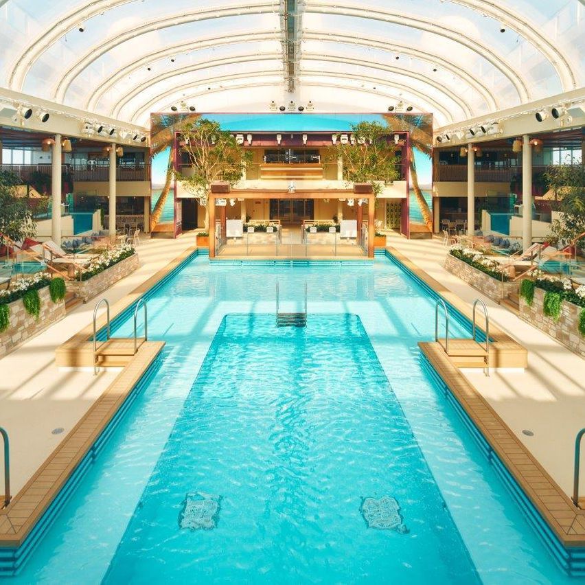 Ein großes Hallenbad mit Glasdach und Sprungbrett auf der linken Seite. Der Pool ist von Liegestühlen und Pflanzen umgeben.