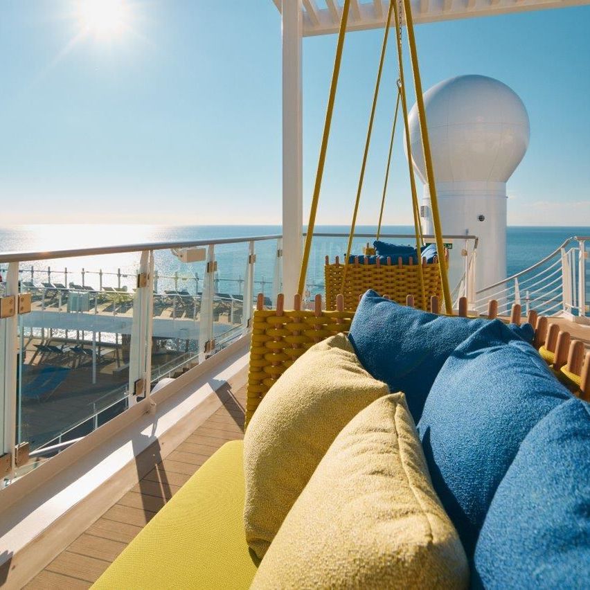 Ein sonnendurchfluteter Deck eines Kreuzfahrtschiffes mit Blick auf den Ozean. Eine Hängematte schwingt von der Decke, und ein Liegestuhl hat blaue und gelbe Kissen.