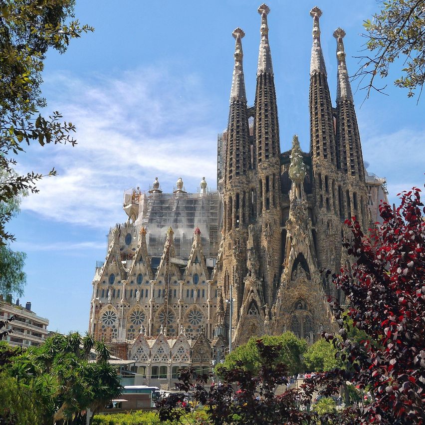 Die Sagrada Familia, eine markante Kathedrale in Barcelona, zeichnet sich durch komplizierte Steinarbeiten und vier hohe Türme aus. Das Gebäude befindet sich teilweise im Bau, umgeben von Bäumen und Pflanzen.
