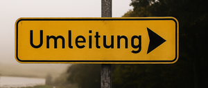 Ein gelbes Schild mit schwarzen Buchstaben und einem Pfeil zeigt nach rechts. Es steht 'Umleitung'. Es ist auf einem Metallpfosten platziert. Dahinter befindet sich eine neblige Landschaft mit einem Grasfeld und Bäumen.