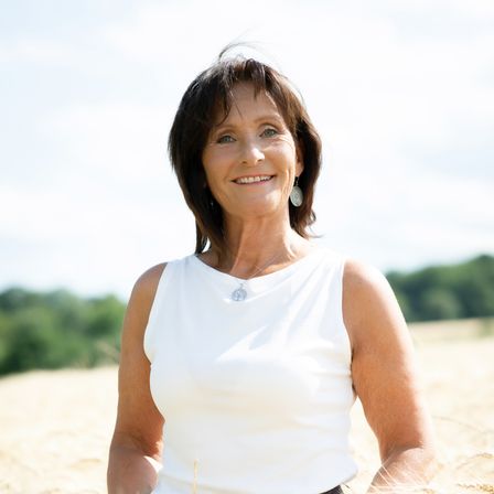 Eine lächelnde Frau in einem weißen ärmellosen Oberteil und einer Halskette sitzt in einem Weizenfeld.