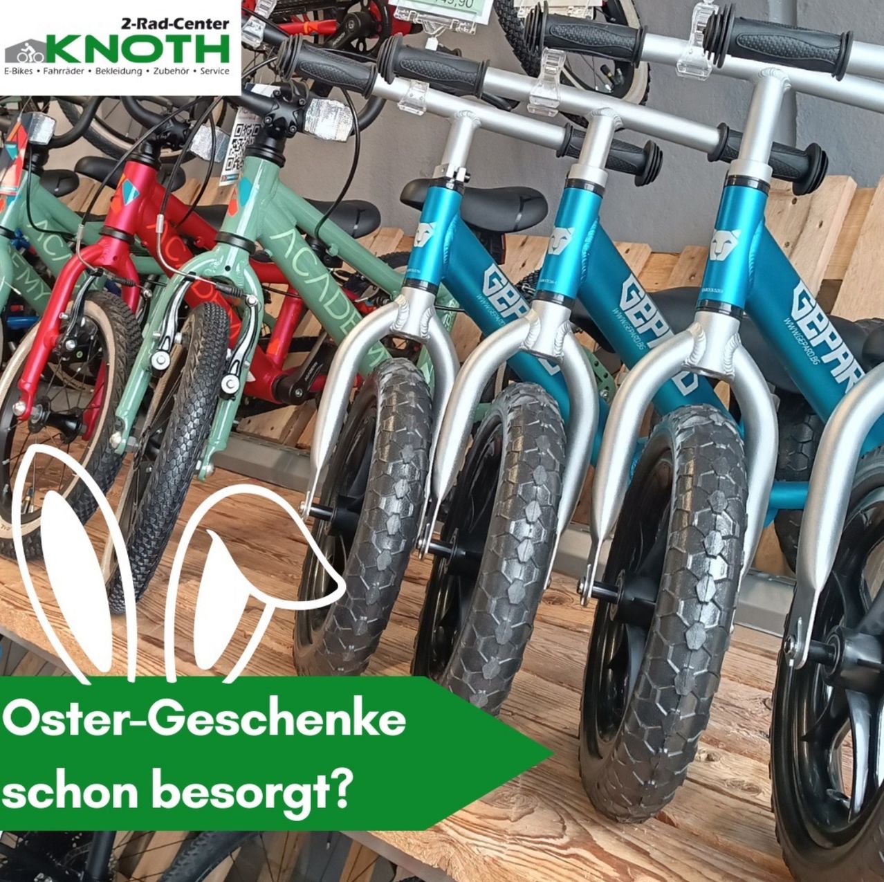 Mehrere Kinderfahrräder sind in einem Geschäft ausgestellt. Sie sind blau, grün und rot. Die Fahrräder stehen auf einer Holzoberfläche. Das Bild hat einen Wasserzeichen mit dem Text 'Oster-Geschenke schon besorgt?' unten links.