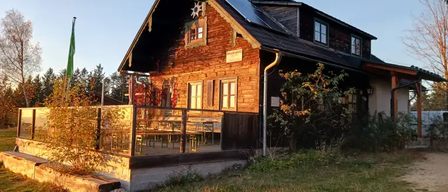 Ein Holzhaus mit Solarpanelen auf dem Dach und einer Veranda mit Tischen und Stühlen. Auf der linken Seite steht eine grüne Fahne.