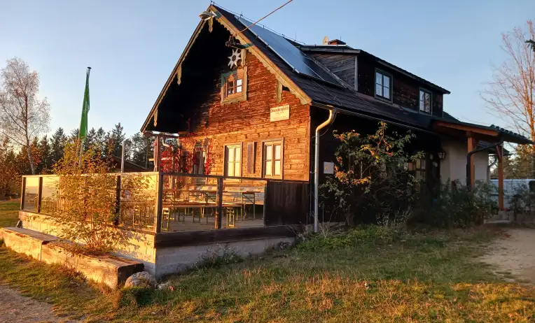 Ein Holzhaus mit Solarpanelen auf dem Dach und einer Veranda mit Tischen und Stühlen. Auf der linken Seite steht eine grüne Fahne.