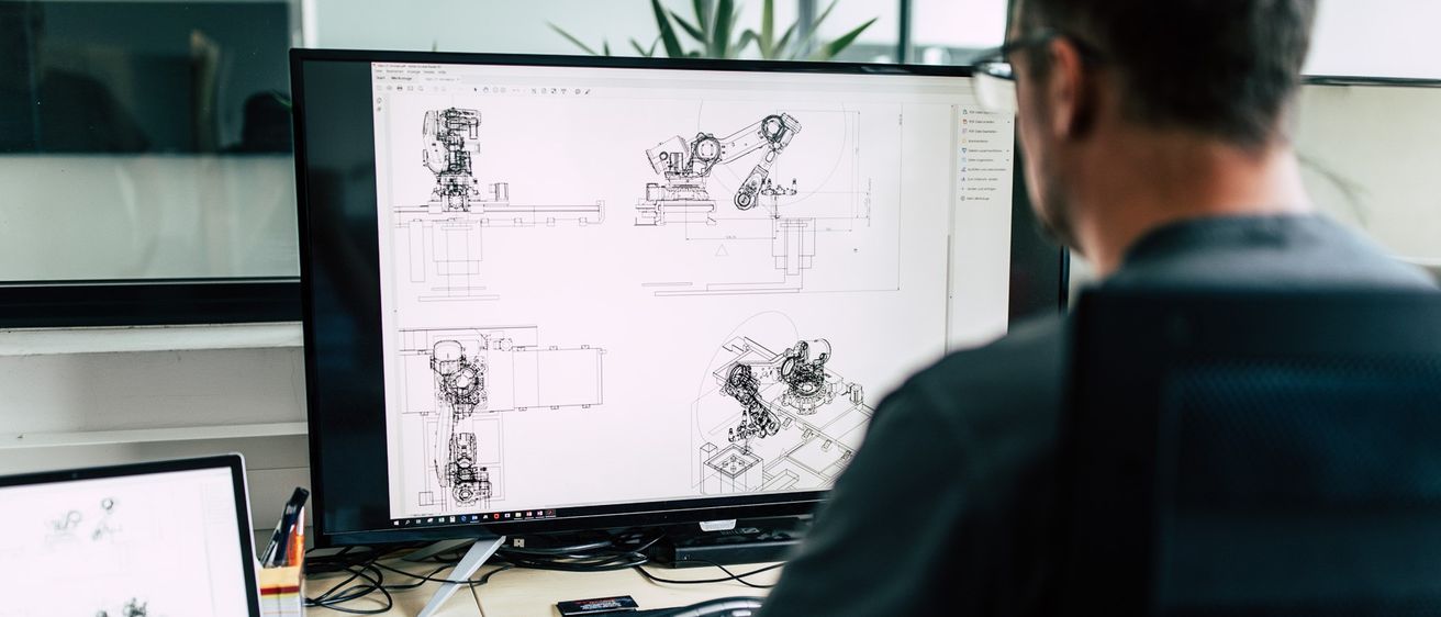 Ein Ingenieur arbeitet an einem großen Monitor, der technische Zeichnungen von Robotermaschinen anzeigt. Er tippt auf einer Tastatur, mit einem Taschenrechner und Dokumenten auf dem Schreibtisch.