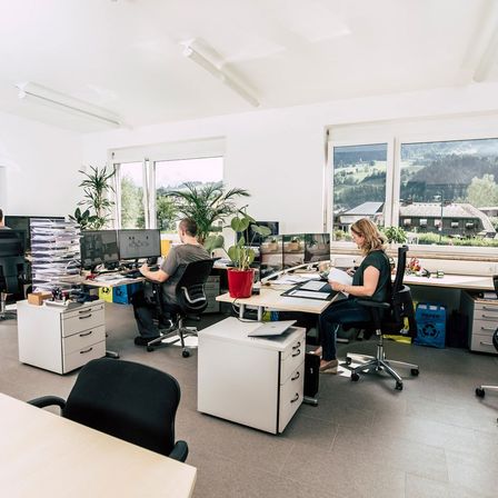 Ein Büroraum mit zwei Mitarbeitern. Ein Mann sitzt an einem Schreibtisch mit mehreren Computerbildschirmen, während eine Frau an einem anderen Schreibtisch einen Papier liest. Pflanzen und Bürozubehör sind auf den Schreibtischen.