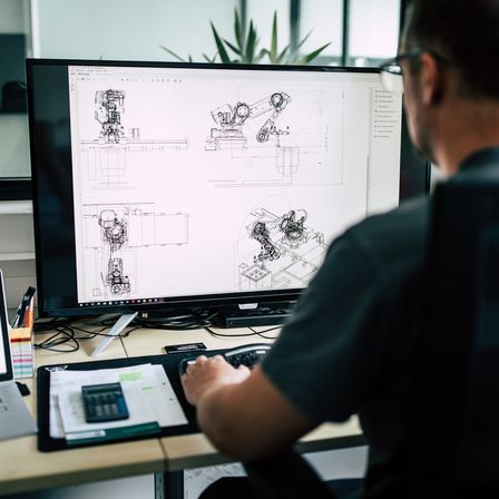 Ein Mann arbeitet an einem Schreibtisch und konzentriert sich auf einen Computerbildschirm mit detaillierten technischen Zeichnungen. Er trägt eine Brille und ein dunkles Hemd.