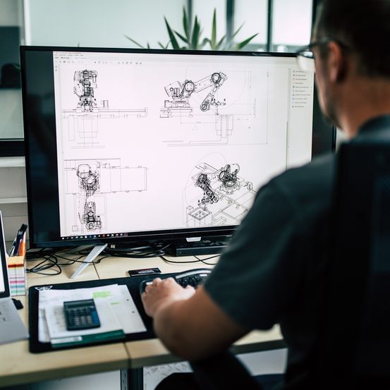 Ein Mann arbeitet an einem Schreibtisch und konzentriert sich auf einen Computerbildschirm mit detaillierten technischen Zeichnungen. Er trägt eine Brille und ein dunkles Hemd.