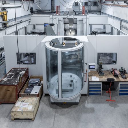 Eine Industrieanlage mit einer großen kreisförmigen Maschine und Glasgehäuse. In der Nähe befinden sich Lagerboxen, ein Werkzeugtisch mit Werkzeugen und verschiedene Geräte. Die Umgebung ist geräumig und gut beleuchtet.