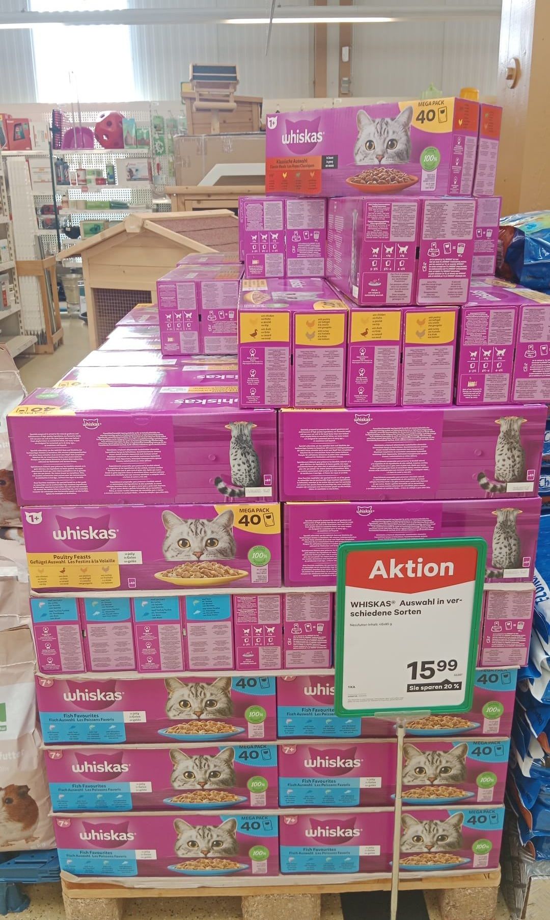 Ein Stapel pinker und gelber Kartons mit Katzenfutter, beschriftet mit 'Whiskas', mit einem Bild einer Katze auf der Vorderseite. Die Kartons sind in einer Ladenumgebung angeordnet.