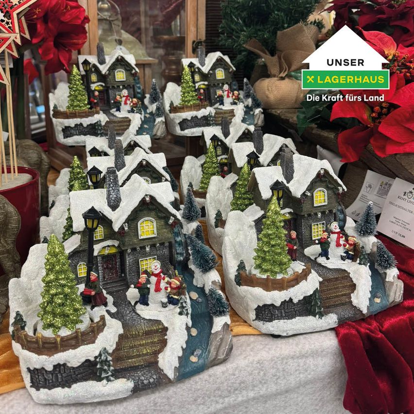 A Christmas village scene with several small houses covered in snow, adorned with trees and festive decorations, is displayed on a table with red cloth.