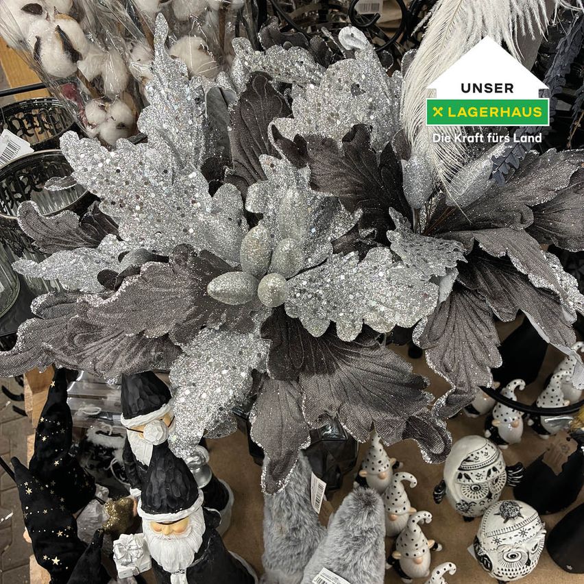 A silver and gray decorative flower arrangement is showcased. It is placed on a brown surface. Various figurines and other objects are around it. A white and green logo reads 'Unser Lagerhaus'.