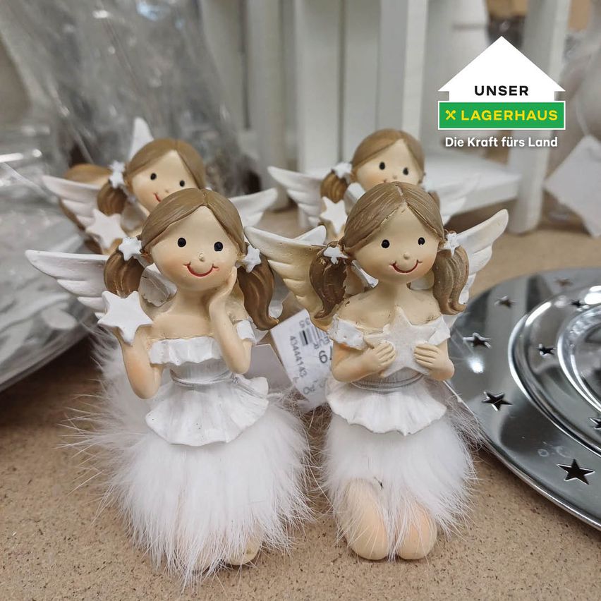 A group of white angel figurines are displayed on a table, each holding a star and smiling. They have white feather skirts and white wings. Behind them is a blurry background with a plate and a tag.