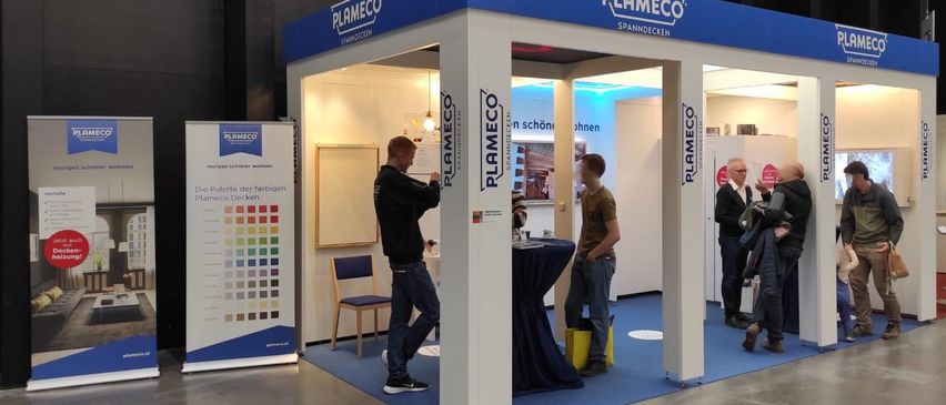 Ein blauer und weißer Stand mit dem Wort Plameco ist in einem Raum aufgebaut. Drinnen sind zwei Männer, die etwas betrachten. Ein Tisch mit einem blauen Tuch steht vor ihnen. Zwei Personen stehen außerhalb des Standes.