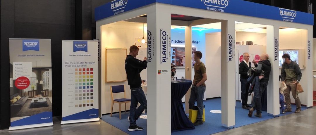 Ein blauer und weißer Stand mit dem Wort Plameco ist in einem Raum aufgebaut. Drinnen sind zwei Männer, die etwas betrachten. Ein Tisch mit einem blauen Tuch steht vor ihnen. Zwei Personen stehen außerhalb des Standes.