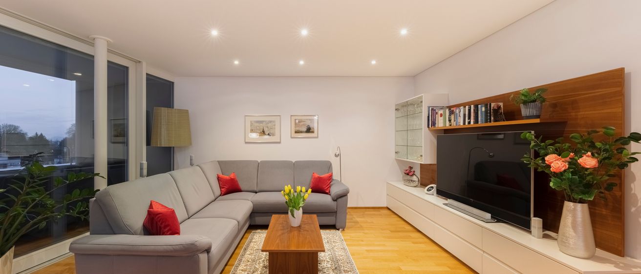 Ein modernes Wohnzimmer mit einem großen grauen Ecksofa, roten Kissen, einem Holztisch mit gelben Blumen, einem Flachbildfernseher und einem weißen Entertainment-Center mit Büchern und einem Topf.