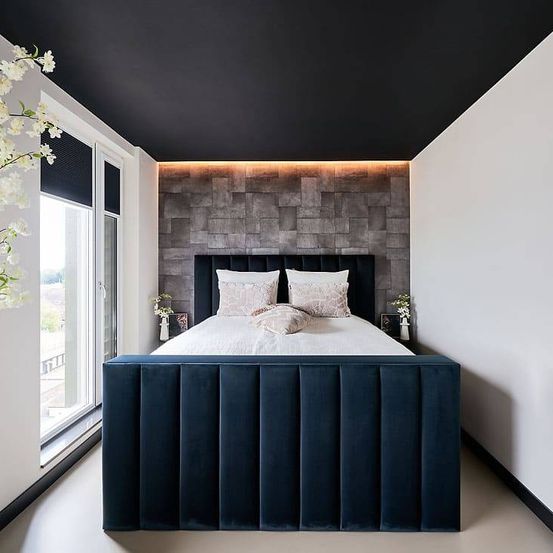 Ein modernes Schlafzimmer mit schwarzer Decke und grauer Steinwand. Ein blaues Bett mit weißen Laken und Kissen steht an der Wand. Auf beiden Seiten des Bettes stehen zwei Vasen mit weißen Blumen.
