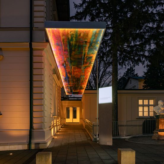 Beleuchteter Gebäudeeingang mit farbenfroher Decke, einer Büste auf einem Sockel und Bäumen bei Nacht.