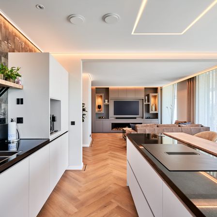 Moderner offener Wohnbereich mit großer Küche und Wohnbereich mit Kamin und Fernseher. Holzböden und weiße Schränke mit schwarzer Arbeitsplatte.