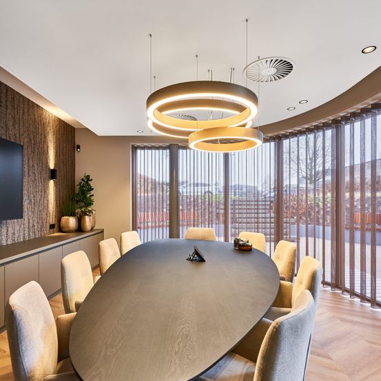 Ein leerer moderner Konferenzraum mit einem großen ovalen Tisch, gepolsterten Stühlen, Holzböden und großen Fenstern. Es gibt zwei runde Pendelleuchten und einen Fernseher an der Wand. Draußen gibt es einen Blick auf Bäume und ein Gebäude.