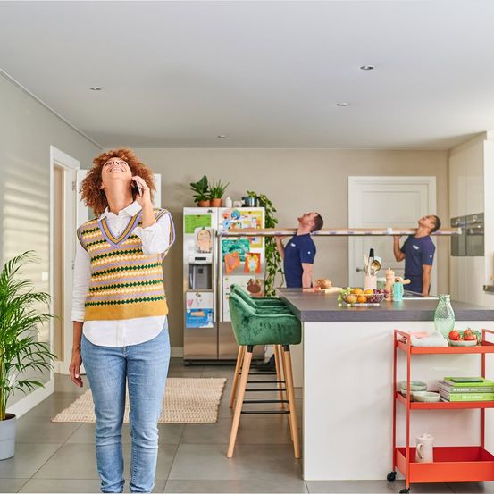 Eine Frau telefoniert in der Küche, mit zwei Männern, die hinter einer Theke mit einer Bar-Höhe-Insel und Pflanzen und einem Kühlschrank stehen.