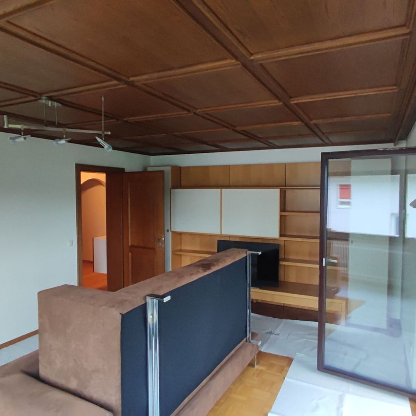 Ein Wohnzimmer mit Holzmöbeln, einem Sofa, einem Regal und einem Fernseher. Die Decke hat Holzpaneele und es gibt eine Glastür. Der Raum ist sauber und gut beleuchtet.