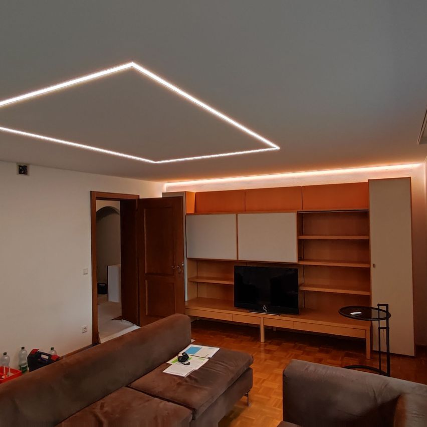 Ein modernes Wohnzimmer mit einer braunen Couch, einem Fernseher auf einem Schrank und einem kleinen Tisch mit einem Notizbuch darauf. Die Decke hat ein geometrisches Muster mit Lichtern.