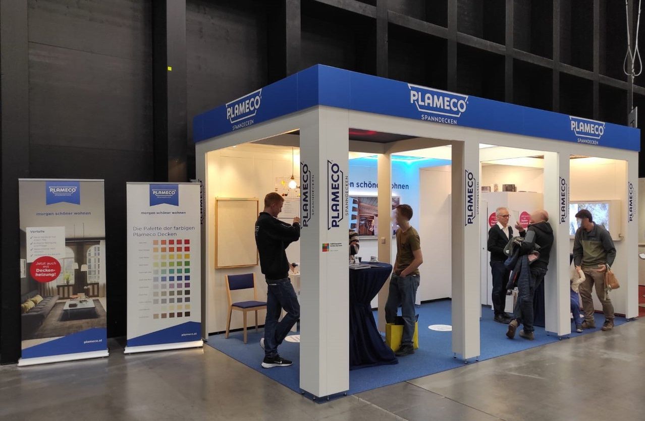A man writes on a board in a Plameco booth, where another man stands with a bag. A third man talks to two others. Plameco branding is on the booth.