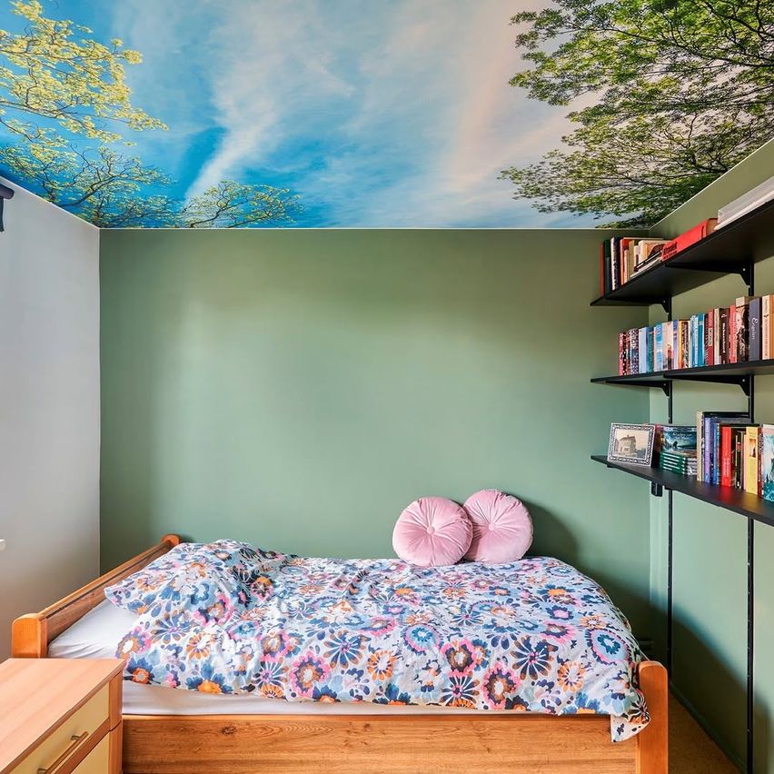 A bedroom with a wooden bed, two pink pillows, and a floral duvet cover. The ceiling features a mural of trees and a blue sky. There is a wooden dresser and a shelf with books.