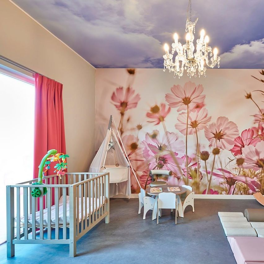 A baby's room with a crib, toys, a table and chairs, and a chandelier on the ceiling. The walls are adorned with floral wallpaper.