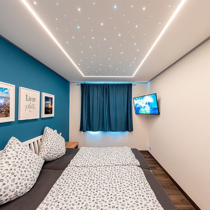 A bedroom with blue walls and a white ceiling with star-like lights. A double bed, pillows, and framed pictures adorn the space. A television is mounted on the wall, and blue curtains cover the window.